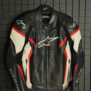 Könnte beinhalten: Eine schwarze, weiße und rote Motorradjacke aus Leder mit dem Alpinestars-Logo auf Brust, Schultern und Ärmeln. Die Jacke verfügt über einen durchgehenden Frontreißverschluss, einen Stehkragen mit Druckknopfverschluss und perforierte Ledereinsätze.
