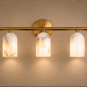 May include: A wall-mounted light fixture featuring a horizontal brass-toned metal bar attached to a circular backplate. Three cylindrical, dome-topped shades with a white and beige marbled pattern hang from the bar, each emitting a warm glow.