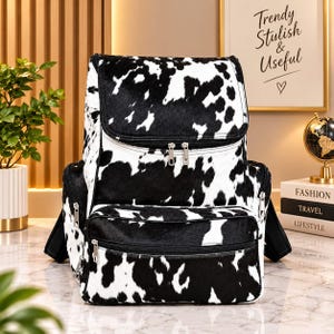 May include: A backpack featuring a black and white cow print pattern. The bag includes a large main compartment with a flap closure, a front zippered pocket, and two side zippered pockets, all equipped with silver-toned metal hardware.