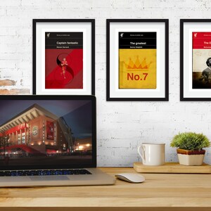 May include: Three framed prints with black frames and white mats. The prints are of a red, white, and black design with the text "Captain Fantastic", "The Greatest", and "The Egyptian King". The prints are hanging on a white brick wall. A laptop computer is on a wooden desk in front of the prints. The laptop screen shows a picture of a stadium at night.