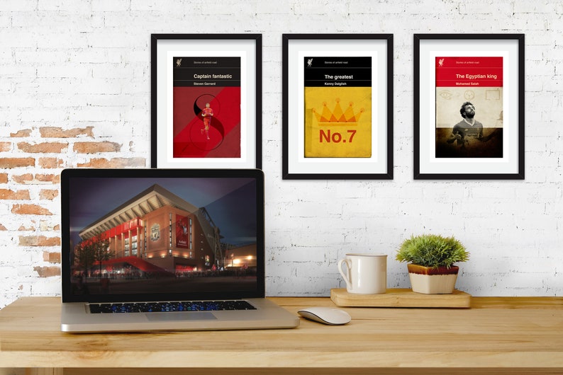 May include: Three framed prints with black frames and white mats. The prints are of a red, white, and black design with the text "Captain Fantastic", "The Greatest", and "The Egyptian King". The prints are hanging on a white brick wall. A laptop computer is on a wooden desk in front of the prints. The laptop screen shows a picture of a stadium at night.