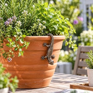 Può includere: Un vaso da fiori in terracotta con un esterno strutturato e scanalato, caratterizzato da un ornamento a forma di lucertola in metallo color bronzo scuro agganciato al bordo. Il vaso è riempito di erbe verdi, tra cui rosmarino e piccoli fiori bianchi e viola.