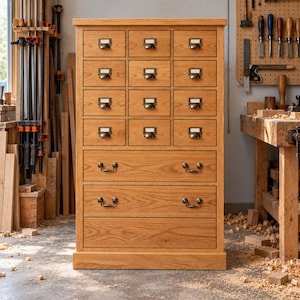 May include: A tall wooden cabinet featuring twelve small drawers with brass label holders and three large bottom drawers with brass pull handles. The light brown oak unit has a flat top and a sturdy base, standing in a workshop environment.