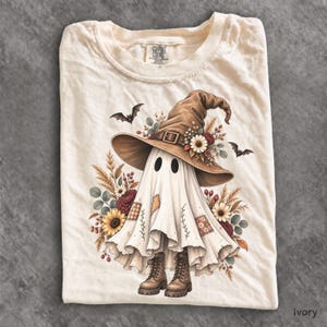 May include: An ivory t-shirt featuring a graphic of a ghost wearing a large brown witch hat adorned with flowers. The ghost has patches, wears brown lace-up boots, and is surrounded by autumn-themed floral arrangements and two small black bats.
