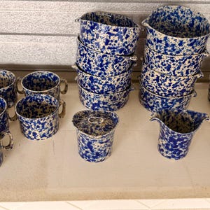 May include: A collection of ceramic dinnerware featuring a blue and white speckled pattern. The set includes multiple handled mugs, stacked bowls, a lidded container, a pitcher, and two stacks of round plates, all sharing a consistent mottled design.