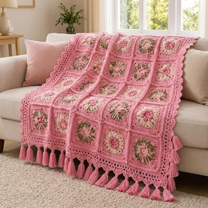 May include: A handmade crochet throw blanket featuring a repeating pattern of square motifs in shades of pink, cream, and sage green. The blanket is finished with a decorative scalloped border and long pink yarn tassels along the bottom edge.