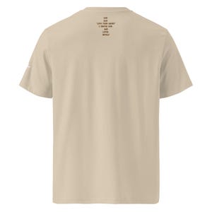 Può includere: Una t-shirt beige a maniche corte con testo marrone scuro stampato sulla parte superiore della schiena che recita: GOD SAID LOVE YOUR ENEMY I OBEYED HIM AND LOVED MYSELF. La maglia presenta un girocollo e un piccolo logo bianco sulla manica sinistra.