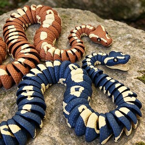 May include: Two articulated snake toys with segmented bodies. One snake is brown with cream-colored patches, and the other is dark blue with cream-colored patches. Both feature textured scales, defined heads with visible eyes, and a flexible design.