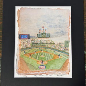 Puede incluir: Una acuarela de Wrigley Field que muestra un partido de béisbol, un marcador con banderas y un letrero que dice 'Cubs 100 Years'. La obra se titula 'My View Of Wrigley Field', tiene fecha del 29 de agosto de 2026 y está en un marco negro.