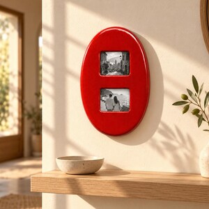 May include: A glossy red oval wall frame featuring two square openings. The top opening displays a black and white photograph of a town street, while the bottom opening contains a black and white photograph of two people standing by the sea.