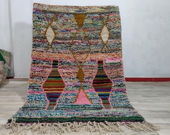 Puede incluir: Una alfombra rectangular con un patrón de rayas horizontales multicolores y una superficie texturizada. El diseño incluye grandes formas de diamante en amarillo mostaza, rosa y blanco, rematadas con flecos de borlas color hueso.