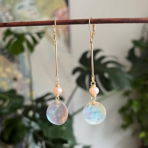 May include: A pair of gold-tone drop earrings featuring a long thin metal bar, a small round peach-colored pearl, and a circular iridescent shell disc at the base. The earrings are suspended from gold-tone lever-back closures.