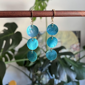 May include: A pair of drop earrings featuring three circular, iridescent turquoise blue discs suspended vertically from gold-toned metal lever-back hooks. Each disc displays a swirling, pearlescent texture that catches the light, creating a layered, dangling effect that hangs from a horizontal wooden bar.