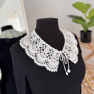 May include: A white crocheted collar featuring an intricate floral pattern with scalloped edges. The collar is secured at the front with a thin crocheted tie that ends in two small, round, matching white crocheted decorative accents.