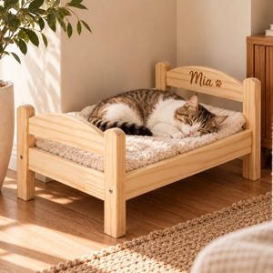 May include: A small wooden pet bed with a light natural finish, featuring a headboard engraved with the name Mia and a paw print icon. A brown and white tabby cat is sleeping on a plush, cream-colored textured cushion inside the frame.