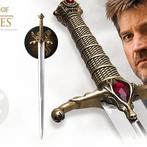Könnte beinhalten: Eine Game of Thrones Schwertnachbildung mit goldenem Griff, einem markanten roten Edelstein an der Parierstange und strukturiertem Griffstück. Die Klinge aus silberfarbenem Stahl ist auf einer runden Holzplatte mit goldenem Löwenemblem montiert.