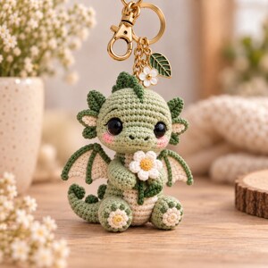 Peut inclure: Un porte-clés dragon vert crocheté avec de grands yeux noirs, des joues roses et de petites ailes. Le dragon tient une fleur blanche au centre jaune. Il est fixé à un anneau en métal doré avec une petite breloque fleur blanche et feuille verte.