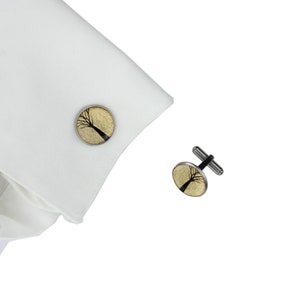 May include: A pair of silver cufflinks with a round, gold-toned design featuring a black silhouette of a tree.