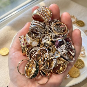 May include: A collection of assorted rings in gold and silver tones, featuring various designs including bands with clear stones, a ring with a large purple gem, a ring with red accents, and a band with green enamel detailing, all held together in a cluster.