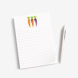 May include: A white notepad featuring a watercolor illustration of three carrots in orange, light orange, and purple at the top. The notepad has horizontal lines for writing, each preceded by a small heart icon, accompanied by a silver ballpoint pen.