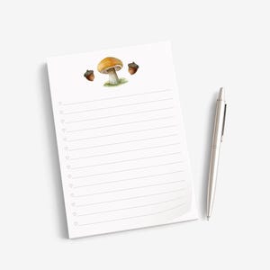 May include: A white notepad with horizontal lines and a small heart icon at the start of each line. The top of the page features a watercolor illustration of a single orange-capped mushroom flanked by two brown acorns. A silver ballpoint pen.