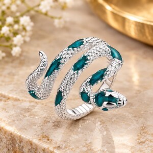 May include: A silver-toned snake ring featuring a textured, scale-like surface and inlaid deep teal enamel accents along the body. The serpent design wraps around the finger, with the head and tail forming the open ends of the coiled band.