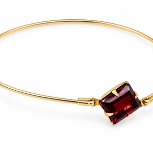 May include: A thin gold bangle bracelet featuring a single rectangular, deep red gemstone set in a four-prong gold setting. The gemstone is positioned as a focal point, connected to the slender, circular gold band by two small hinged joints.
