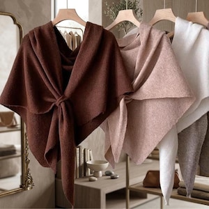 May include: A collection of three soft, textured knit ponchos hanging on wooden hangers. The ponchos are featured in solid colours of dark brown, dusty rose, and white. Each garment has a draped, asymmetrical design with a front tie closure.