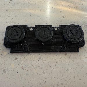 Op de afbeelding: Een zwart 3D-geprint bedieningspaneel met drie cirkelvormige knoppen die horizontaal zijn geplaatst. De linkerknop toont een driehoek die naar boven wijst, de middelste knop een verticale lijn en de rechterknop een driehoek die naar beneden wijst.