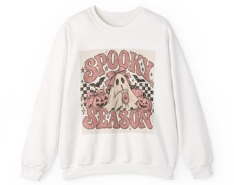 May include: A white crewneck sweatshirt featuring a central graphic with the text SPOOKY SEASON in pink letters. The design includes a ghost wearing a bow, holding a drink, surrounded by pumpkins, bats, stars, and a black and white checkerboard pattern.