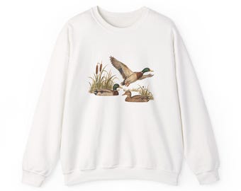 May include: A white long-sleeve crewneck sweatshirt featuring a central graphic illustration of three mallard ducks in a marshy environment with tall reeds and cattails. One duck is in flight while two others are swimming in the water.
