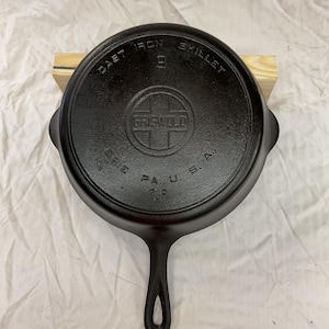 May include: A black cast iron skillet with a long handle. The bottom features embossed text reading CAST IRON SKILLET, a central logo with the word GRISWOLD inside a cross, and ERIE PA. U. S. A. 710 H below the logo. The skillet is round.