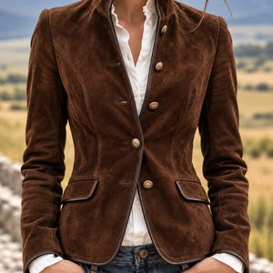 May include: A dark brown suede blazer featuring a high collar, long sleeves, and a front button closure with metallic buttons. The jacket is trimmed with dark brown piping along the edges and includes two front flap pockets, worn over a white shirt.