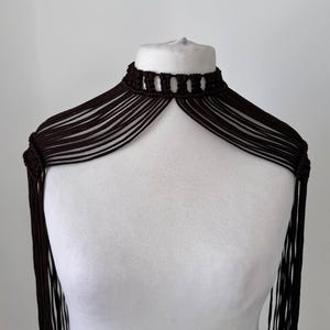 Könnte beinhalten: Ein dunkelbraunes Makramee-Halsband mit hohem Kragen und komplexen Knotendetails. Das Kleidungsstück besteht aus mehreren dünnen, parallelen Kordelsträngen, die über die Schultern fallen und in langen, fließenden Fransen auslaufen.