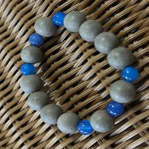 May include: A circular bracelet composed of alternating round beads. The design features matte, textured grey stone beads interspersed with smaller, smooth, translucent bright blue beads arranged in a repeating pattern along the elastic cord.
