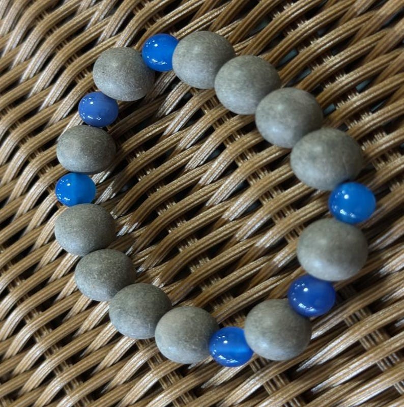 May include: A circular bracelet composed of alternating round beads. The design features matte, textured grey stone beads interspersed with smaller, smooth, translucent bright blue beads arranged in a repeating pattern along the elastic cord.
