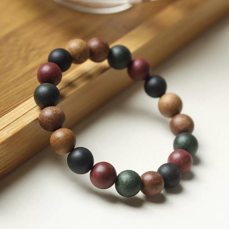 May include: A circular bracelet composed of smooth, matte-finish spherical beads in a repeating pattern of dark green, deep red, black, and natural brown tones. The beads are strung together on an elastic cord, creating a flexible, uniform loop.