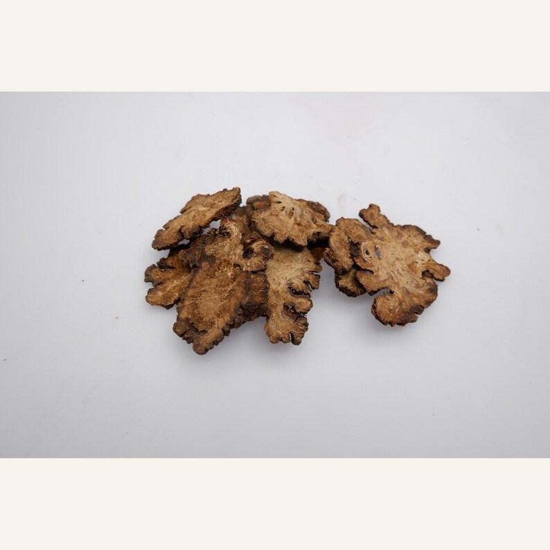 May include: A small cluster of dried, light brown root slices with irregular, jagged edges and a textured, woody surface. The slices are thin and flat, displaying a natural, fibrous appearance with varying shades of tan and dark brown throughout.