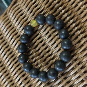 May include: A circular beaded bracelet composed of numerous matte black spherical beads and a single translucent yellow spherical bead. The beads are strung together in a continuous loop, creating a uniform and simple design for the wrist.