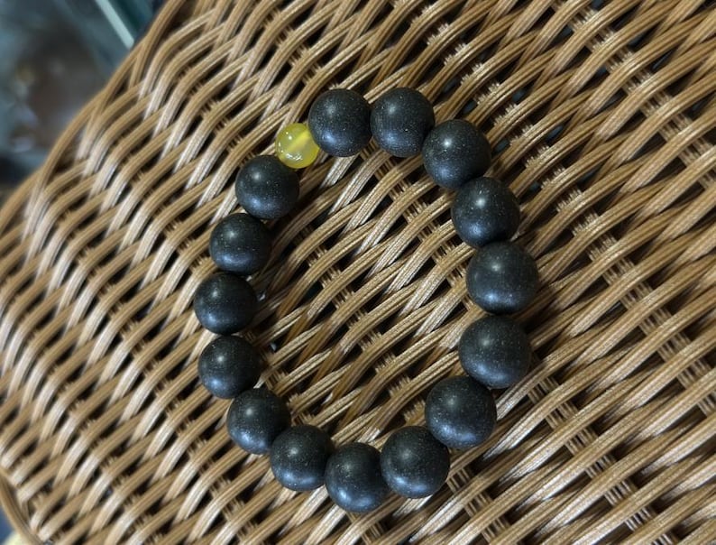 May include: A circular beaded bracelet composed of numerous matte black spherical beads and a single translucent yellow spherical bead. The beads are strung together in a continuous loop, creating a uniform and simple design for the wrist.