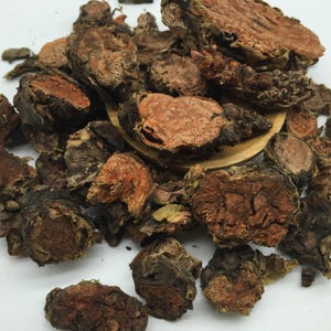 May include: A collection of dried, sliced botanical roots with a rough, dark brown exterior and a fibrous, reddish-orange interior. The pieces vary in size and shape, featuring a dense, woody texture throughout the pile of organic material.