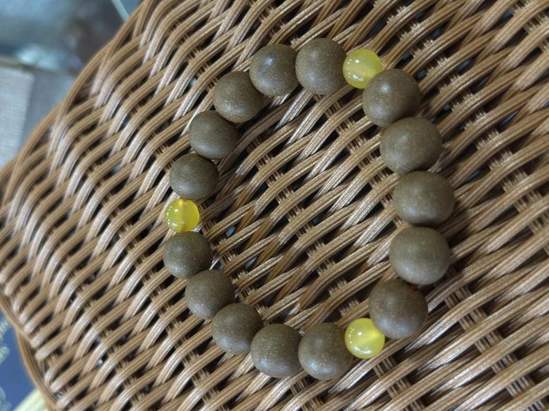 May include: A circular bracelet composed of numerous matte, dark brown, spherical beads interspersed with three smooth, translucent, bright yellow beads. The beads are strung together in a continuous loop, creating a simple and uniform design.