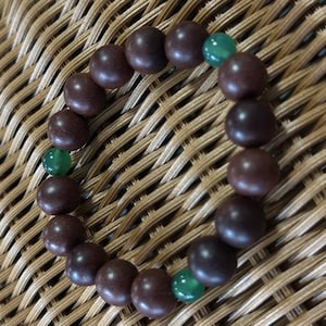 May include: A circular bracelet composed of smooth, dark brown spherical beads interspersed with three smaller, translucent green beads. The beads are strung together in a continuous loop, creating a simple, uniform design with alternating colors.