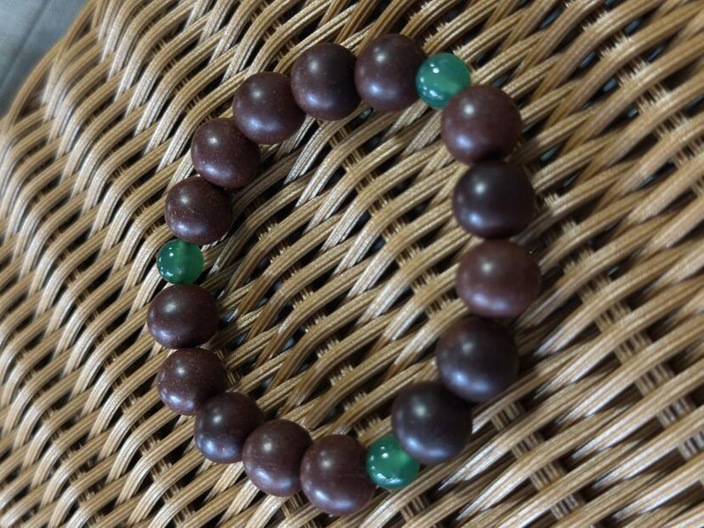 May include: A circular bracelet composed of smooth, dark brown spherical beads interspersed with three smaller, translucent green beads. The beads are strung together in a continuous loop, creating a simple, uniform design with alternating colors.