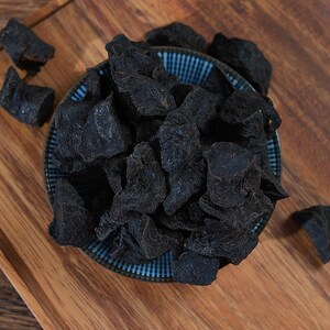 May include: A collection of dark, dried, irregular-shaped pieces of food piled high in a small, round, blue and white checkered ceramic bowl. The pieces have a rough, textured surface and a deep black color, with a few loose pieces scattered nearby.
