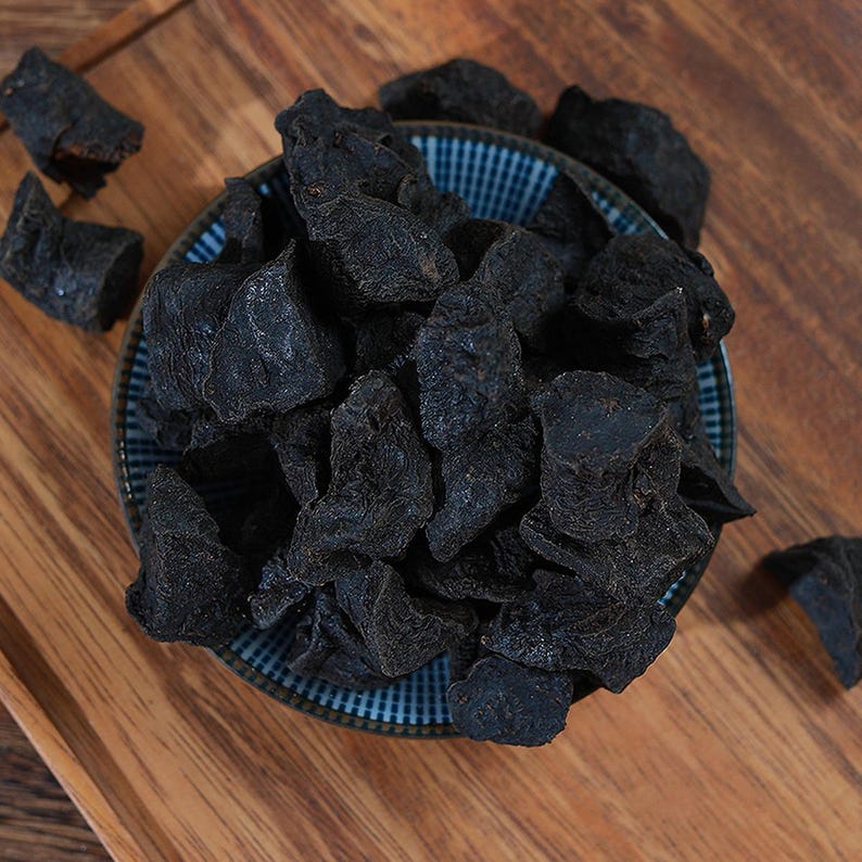 May include: A collection of dark, dried, irregular-shaped pieces of food piled high in a small, round, blue and white checkered ceramic bowl. The pieces have a rough, textured surface and a deep black color, with a few loose pieces scattered nearby.