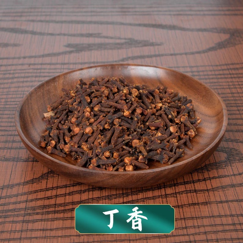 May include: A pile of dried, dark brown whole clove spice buds resting in a shallow, round, polished wooden bowl. The cloves have a distinct nail-like shape with a rounded, textured head. A green rectangular label displays the white text 丁香.