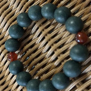 May include: A circular bracelet composed of matte dark green spherical beads arranged in a continuous loop, accented by two smooth, translucent reddish-brown spherical beads positioned on opposite sides of the circular strand.