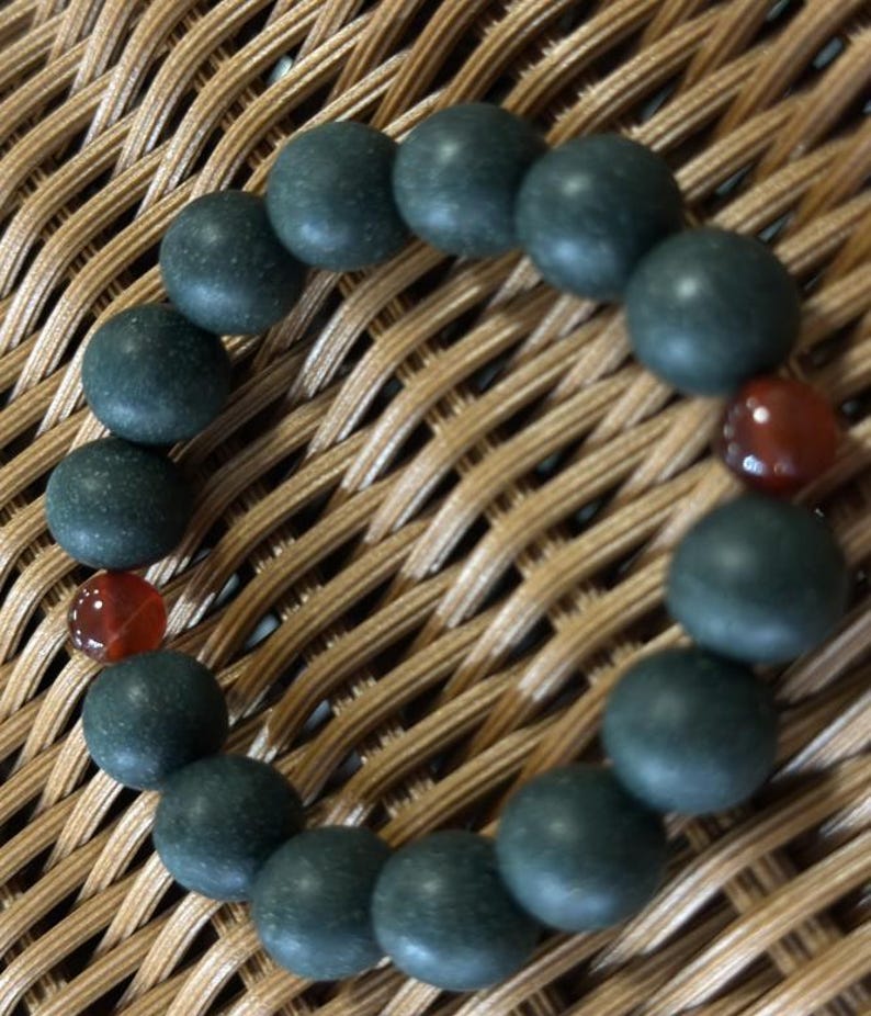 May include: A circular bracelet composed of matte dark green spherical beads arranged in a continuous loop, accented by two smooth, translucent reddish-brown spherical beads positioned on opposite sides of the circular strand.
