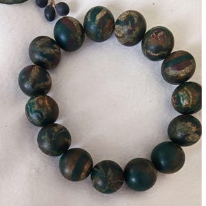 May include: A circular arrangement of fourteen spherical beads, each featuring a mottled surface with deep forest green, earthy brown, and subtle metallic gold-toned swirls. The beads are uniform in size and display a smooth, polished finish.